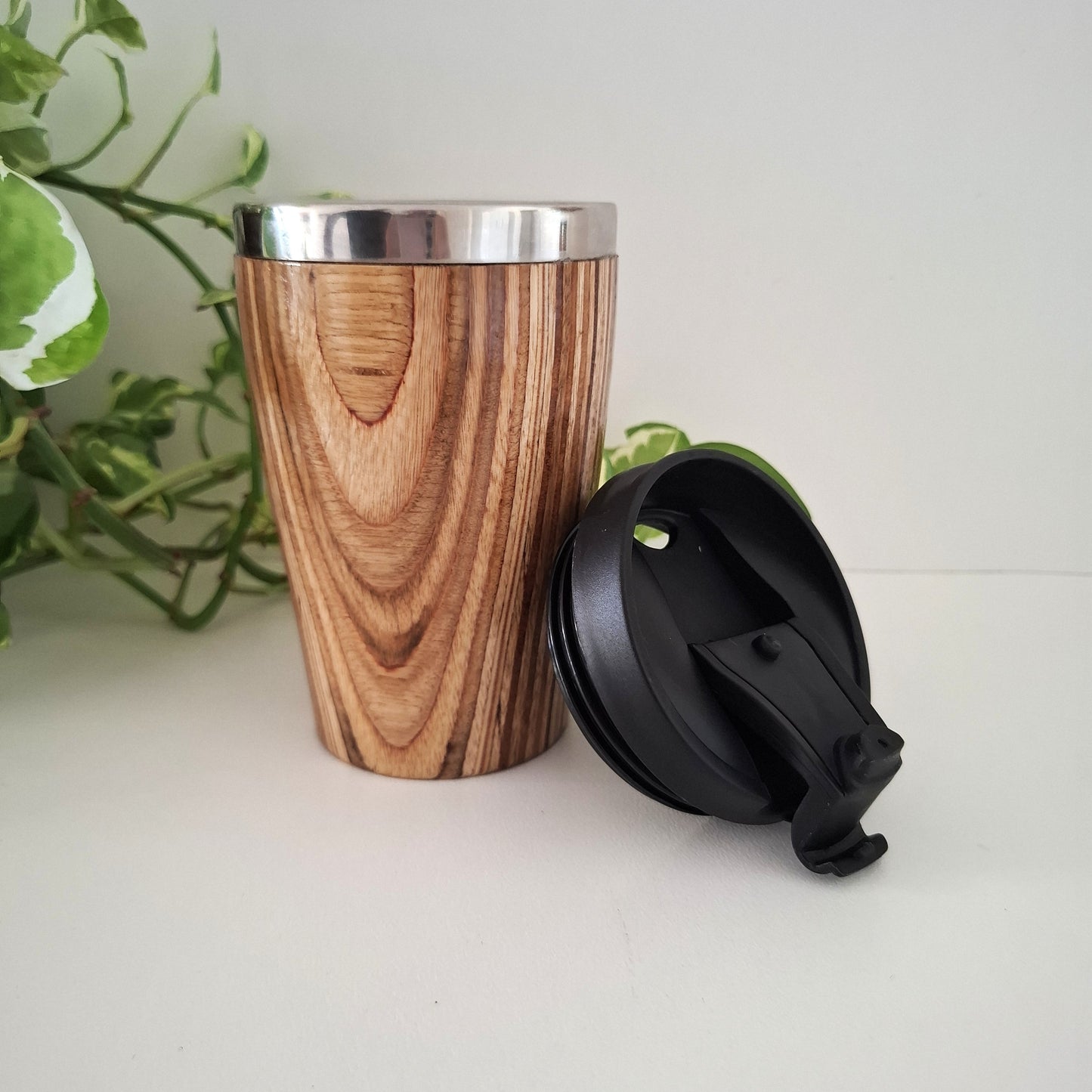 Marnie Ply Wooden travel mug with black lid on a white surface with green plant in the background