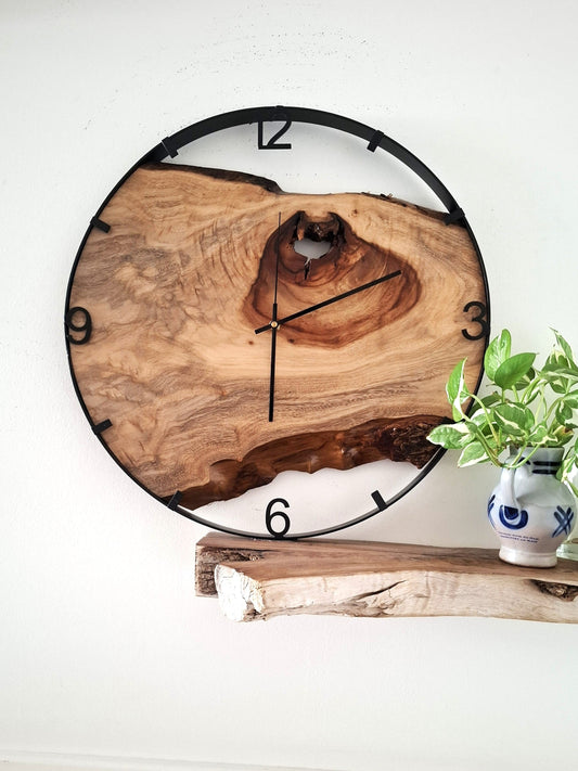 A round wall clock with a natural live-edge wood face and black numerals, mounted on a white wall above a rustic wood shelf with a small potted plant.