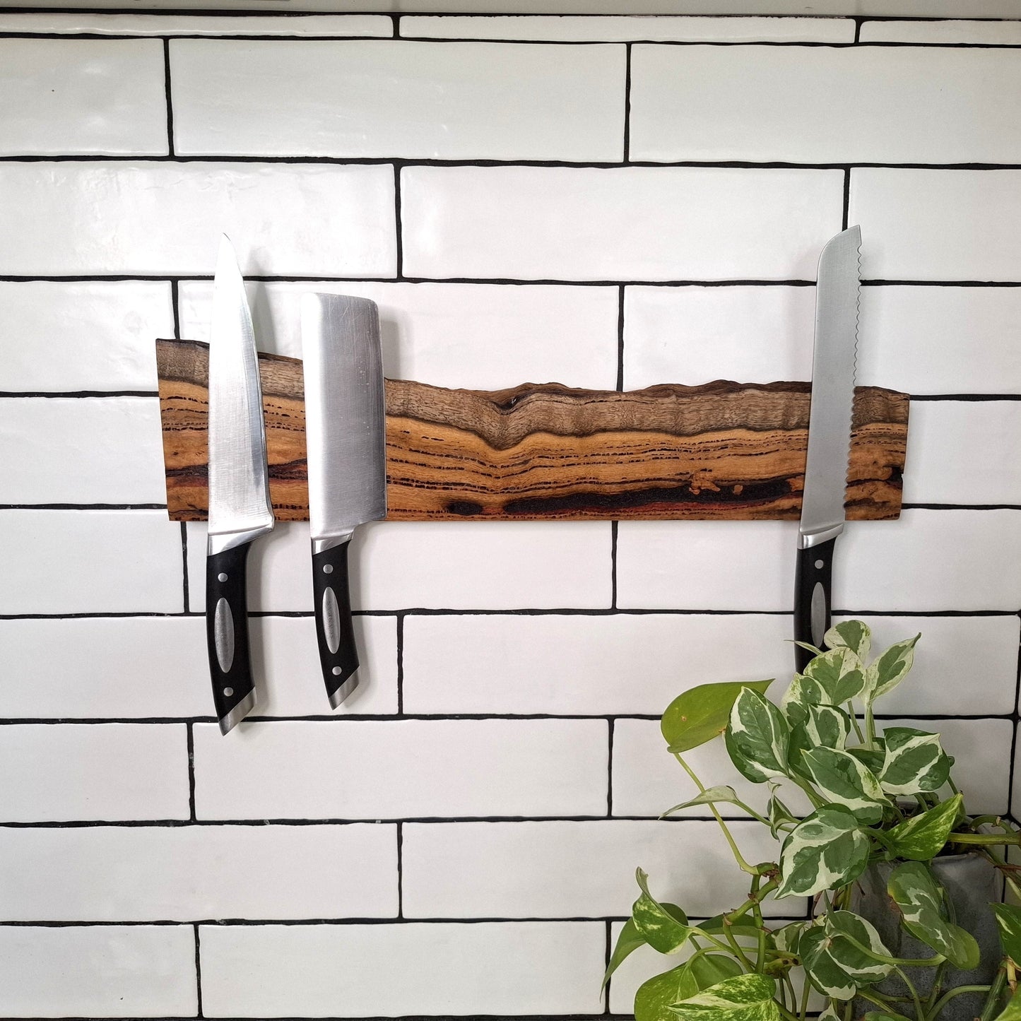 Wooden knife block with knives on a white tiled wall with a plant in the corner.