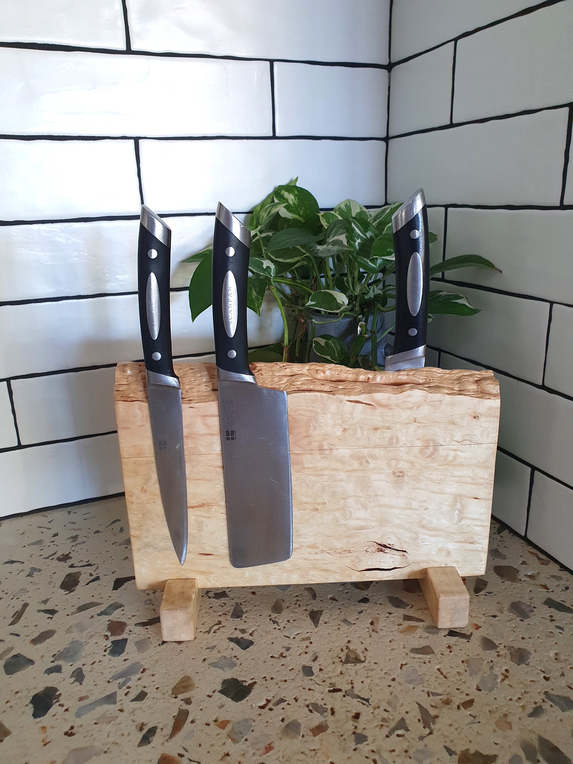 Three knives on a wooden block with a plant in the background against a tiled wall.