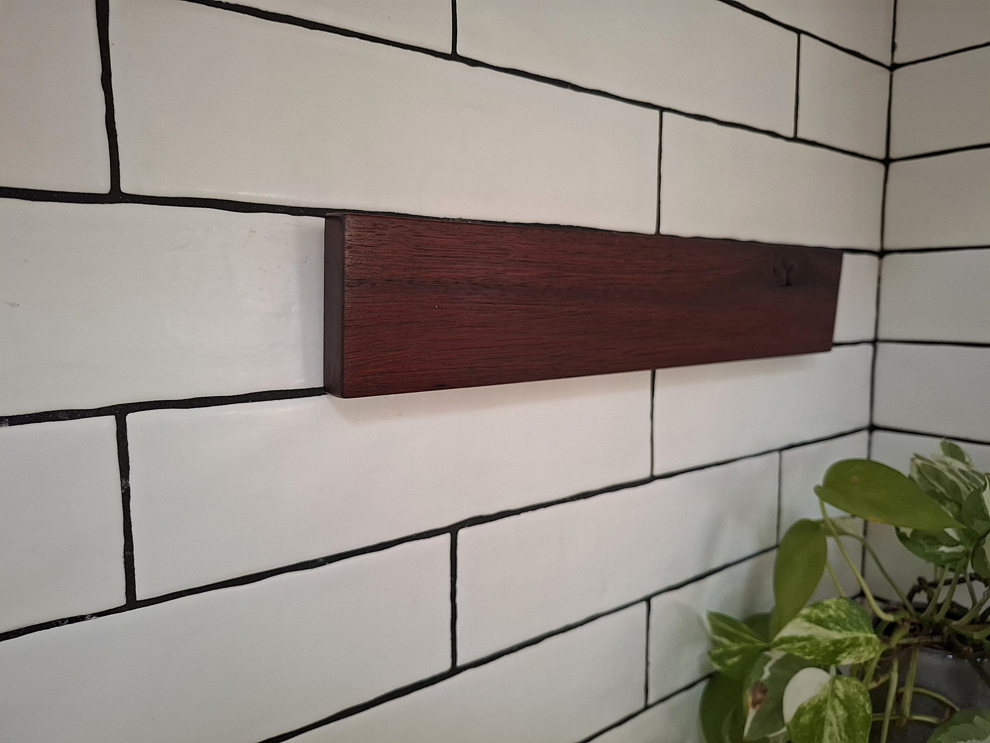 Wooden shelf mounted on a white tiled wall with a plant in the corner.