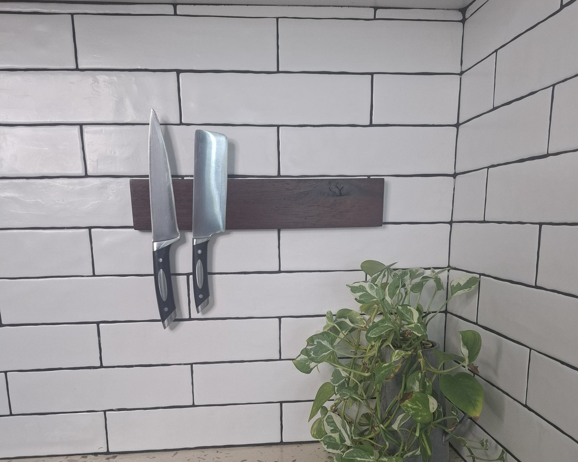 Two knives on a magnetic strip against a tiled wall with a plant in the corner.