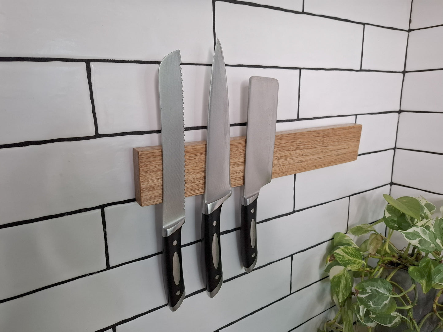 Three knives on a magnetic wooden strip against a tiled wall with a plant in the corner.