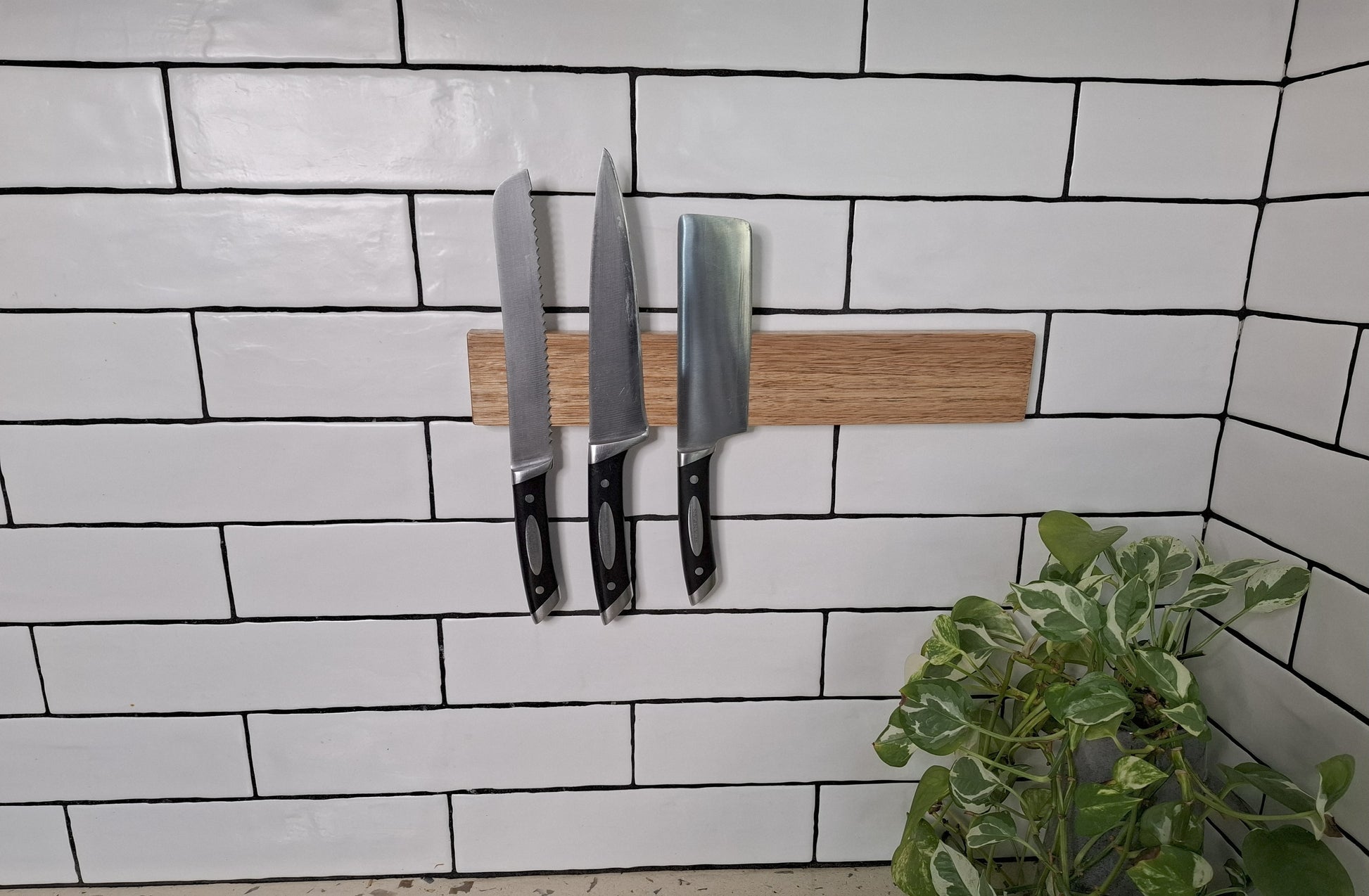 Three knives on a wooden block against a tiled wall with a plant in the foreground.