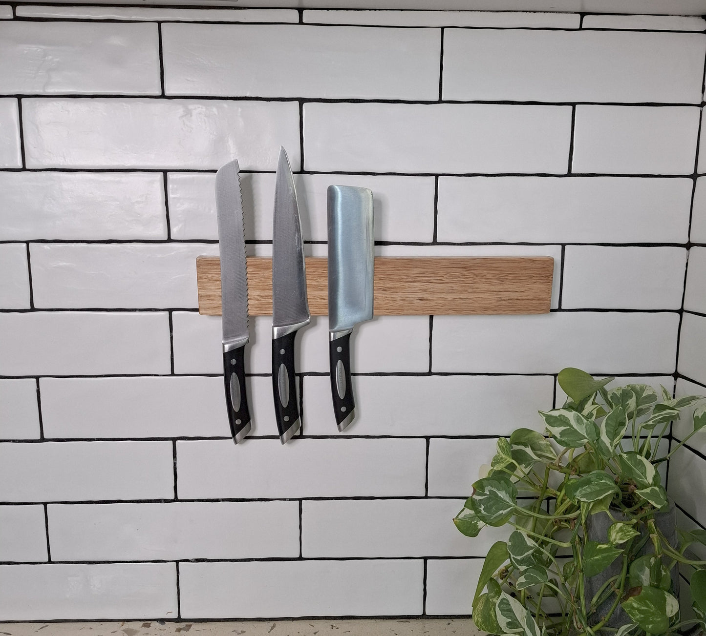Three knives on a magnetic knife holder against a tiled wall with a plant in the corner.