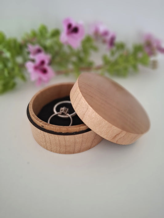 Wooden ring box with a ring inside, placed on a light surface with blurred flowers in the background.