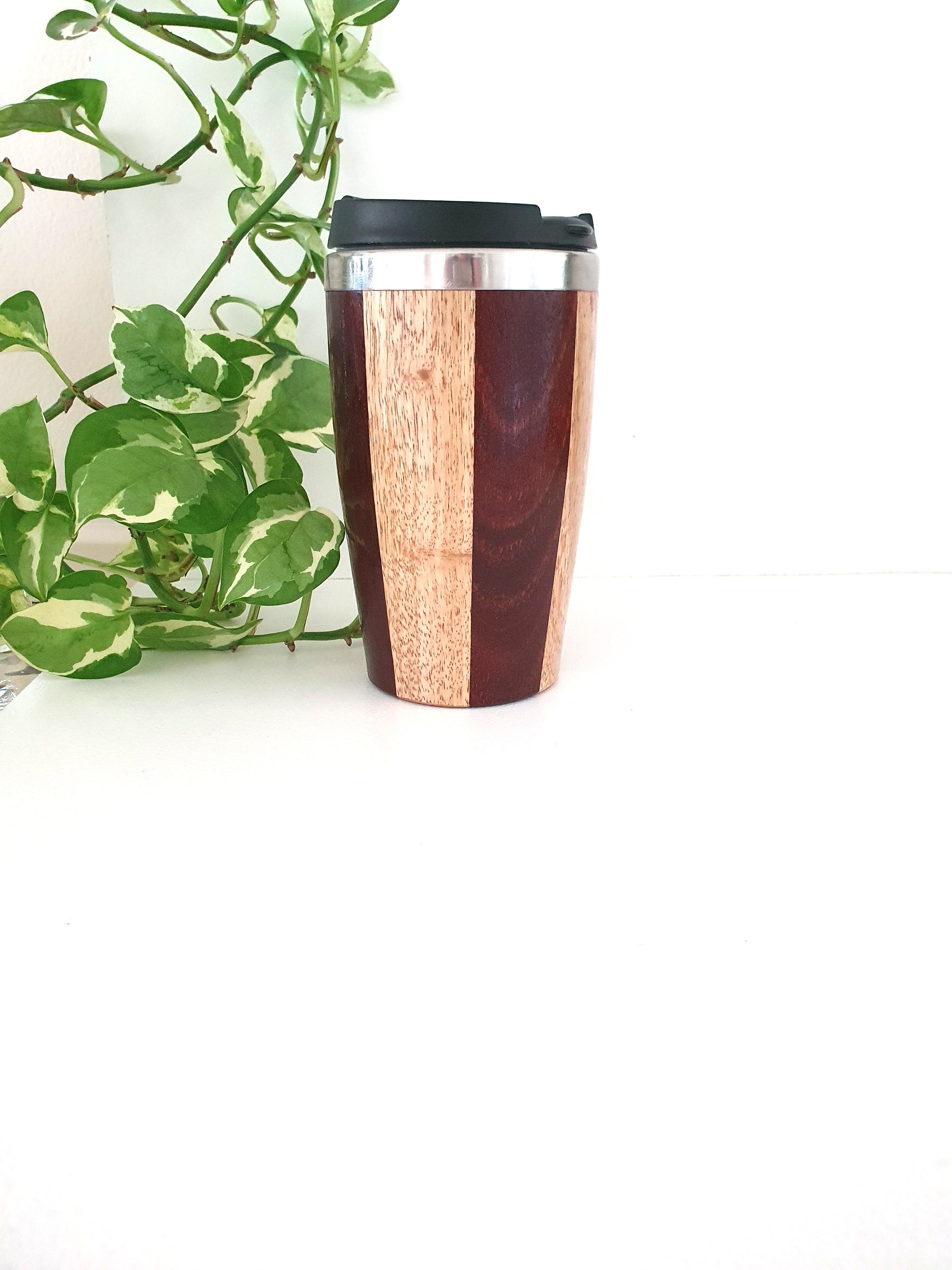 Wooden travel mug with a black lid on a white surface with a green plant in the background