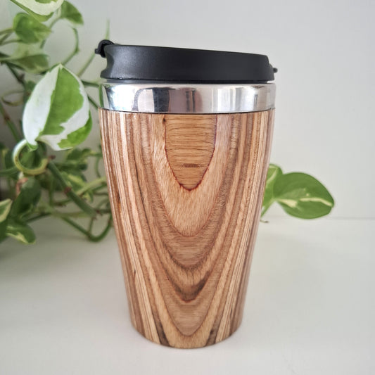Marnie Ply Wooden travel mug with black lid on a white surface with green plants in the background