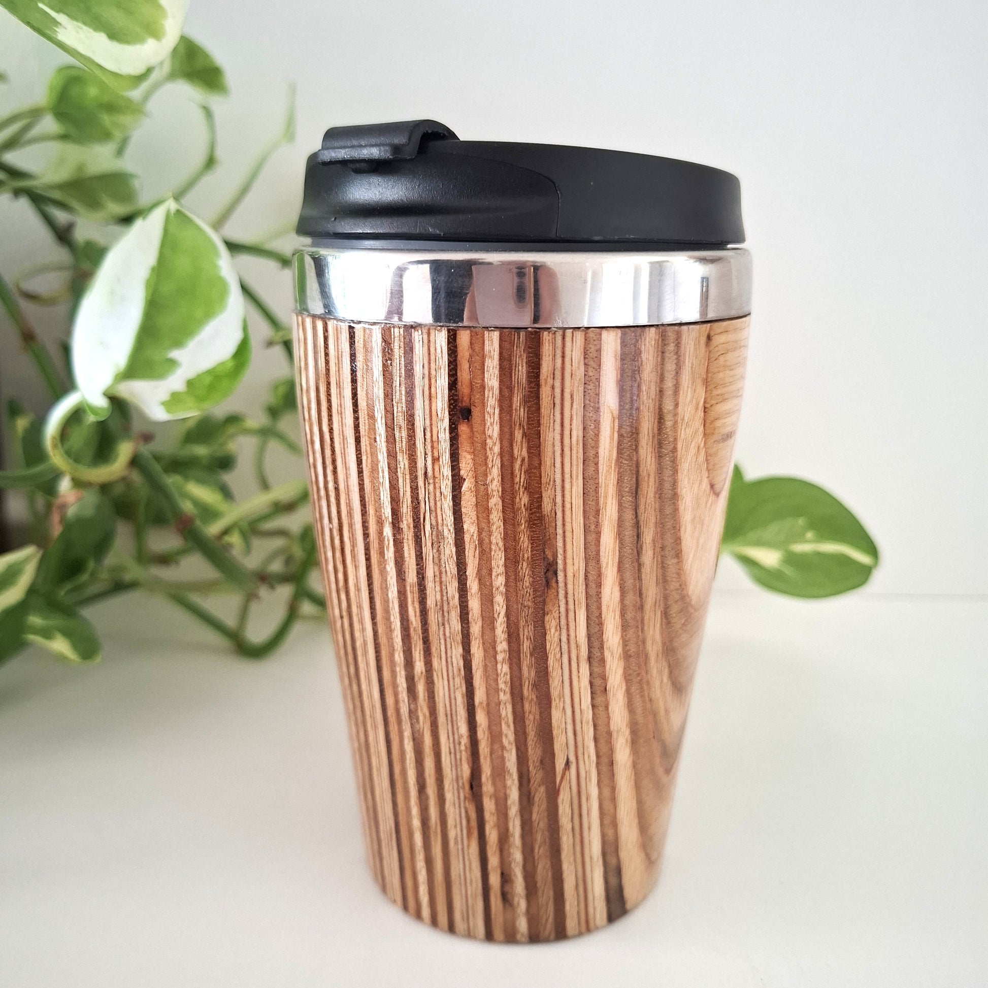 Marnie Ply Wooden travel mug with black lid on a white surface with green plant in the background