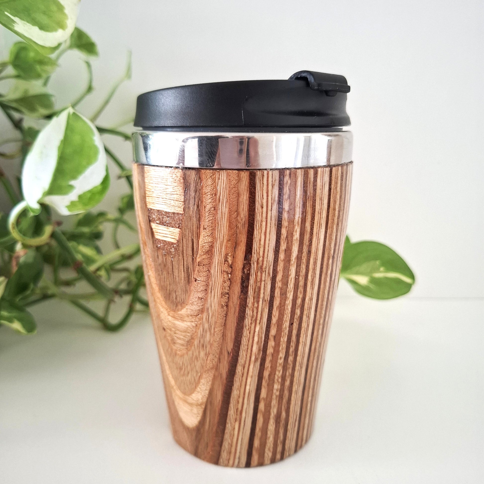 Wooden travel mug with black lid on a white surface with green leaves in the background