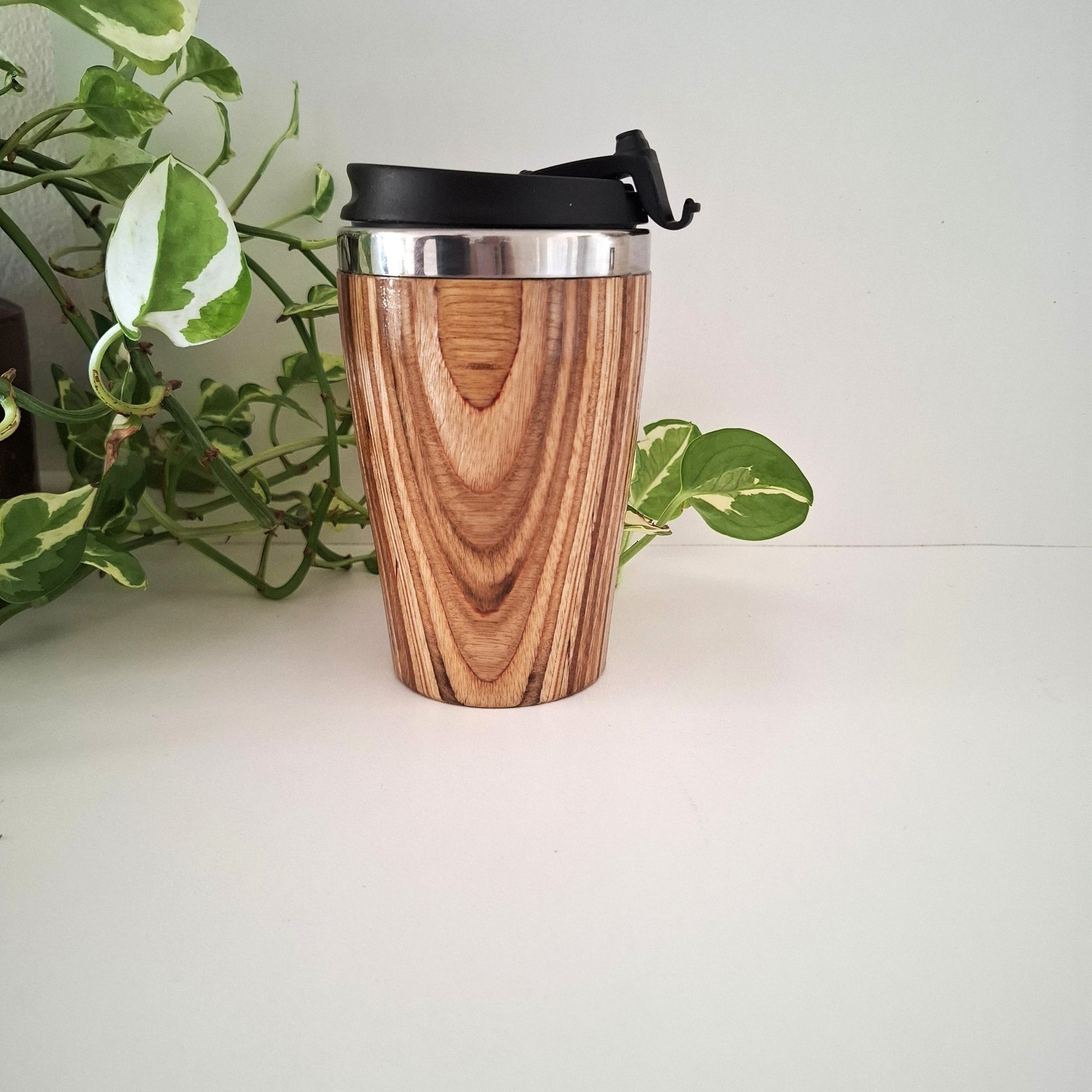 Marnie Ply Wooden travel mug with black lid on a white surface with green plant in the background