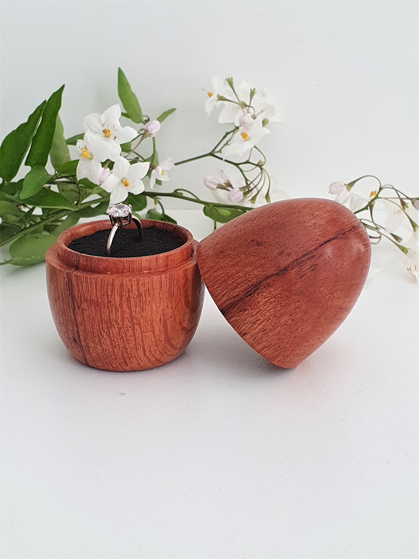 Wooden ring box with a ring inside, surrounded by green leaves and white flowers on a light background