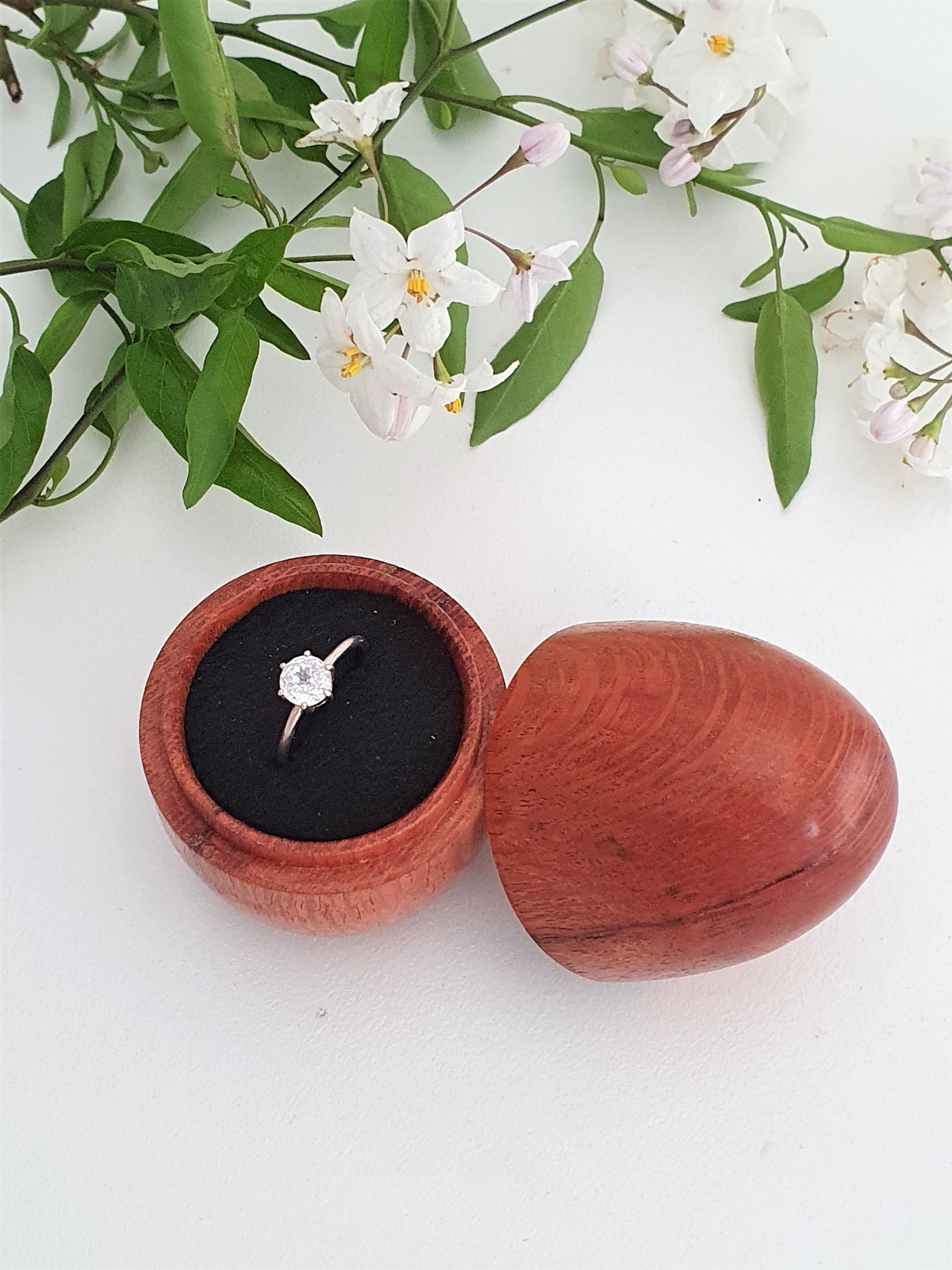 Wooden ring box with a ring inside, surrounded by green leaves and white flowers on a white background