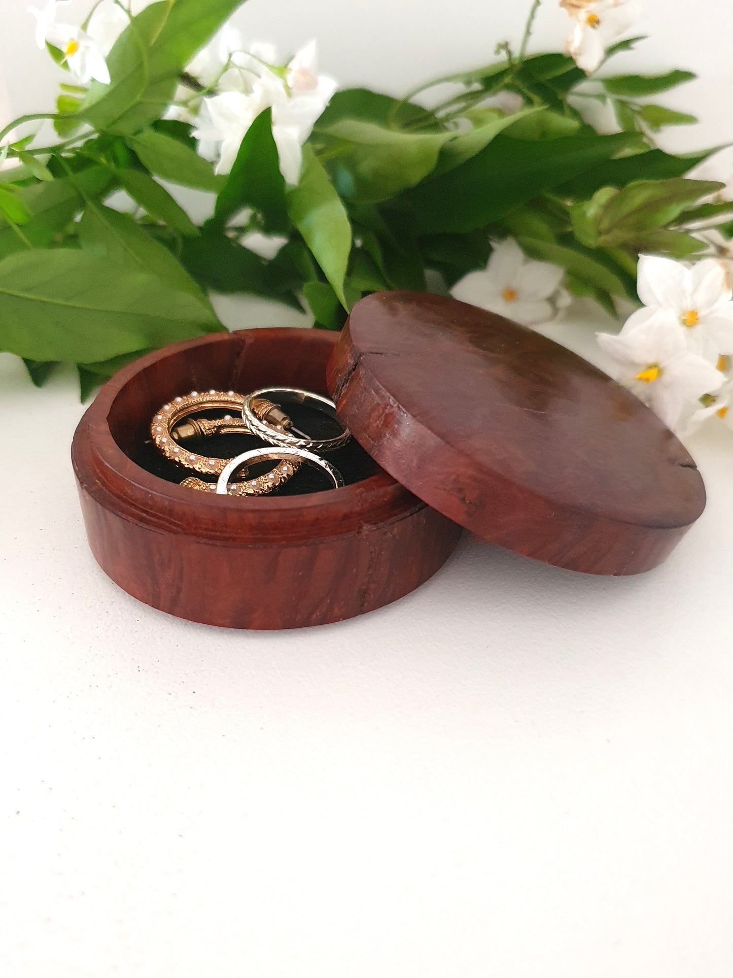 Wooden jewelry box with rings inside, surrounded by green leaves and white flowers on a white background