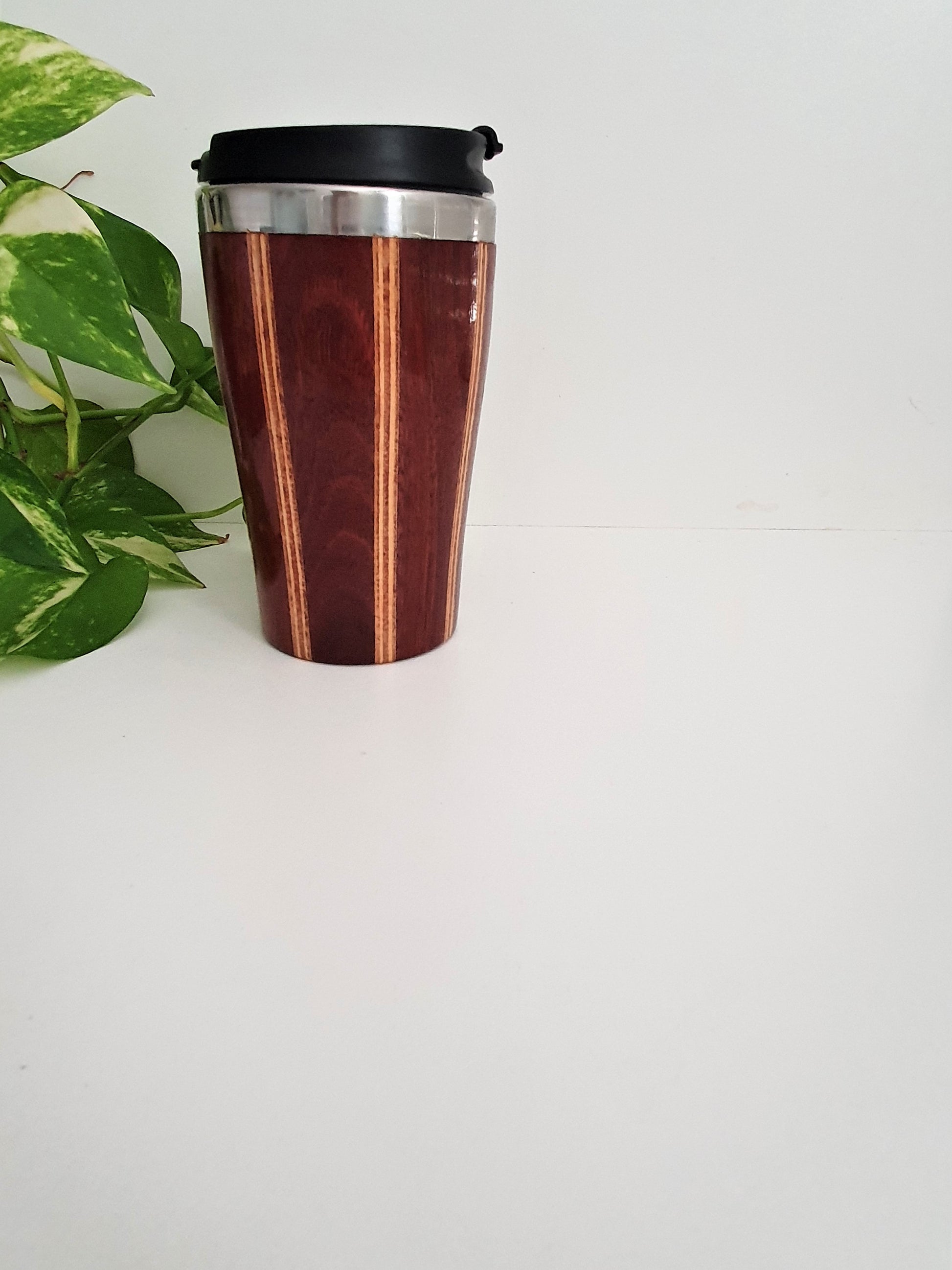 Wooden travel mug with black lid on a white surface with a green plant in the corner.