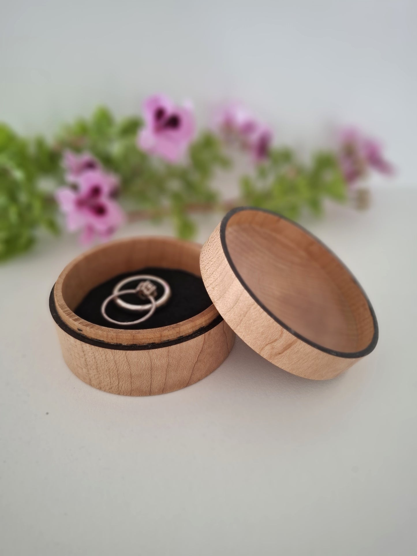 Wooden ring box with a ring inside, placed on a light surface with blurred flowers in the background.